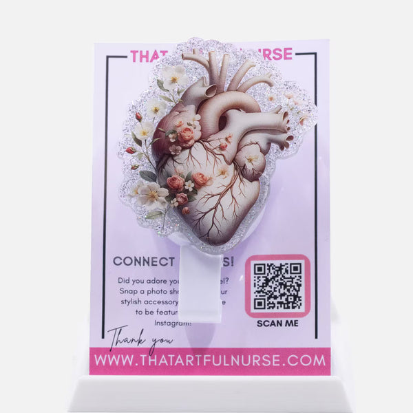 Badge reel featuring an anatomical heart illustration with floral accents and glitter outline, displayed on That Artful Nurse packaging.
