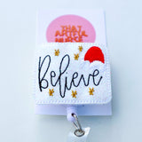 “Believe” Christmas badge reel with gold stars and red Santa hat on a glittery white background.