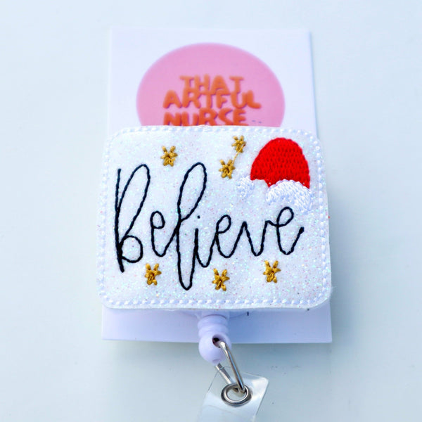 “Believe” Christmas badge reel with gold stars and red Santa hat on a glittery white background.