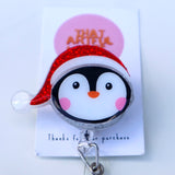 Acrylic Christmas penguin badge reel with red glitter Santa hat and rosy cheeks.
