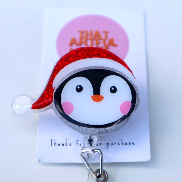 Acrylic Christmas penguin badge reel with red glitter Santa hat and rosy cheeks.