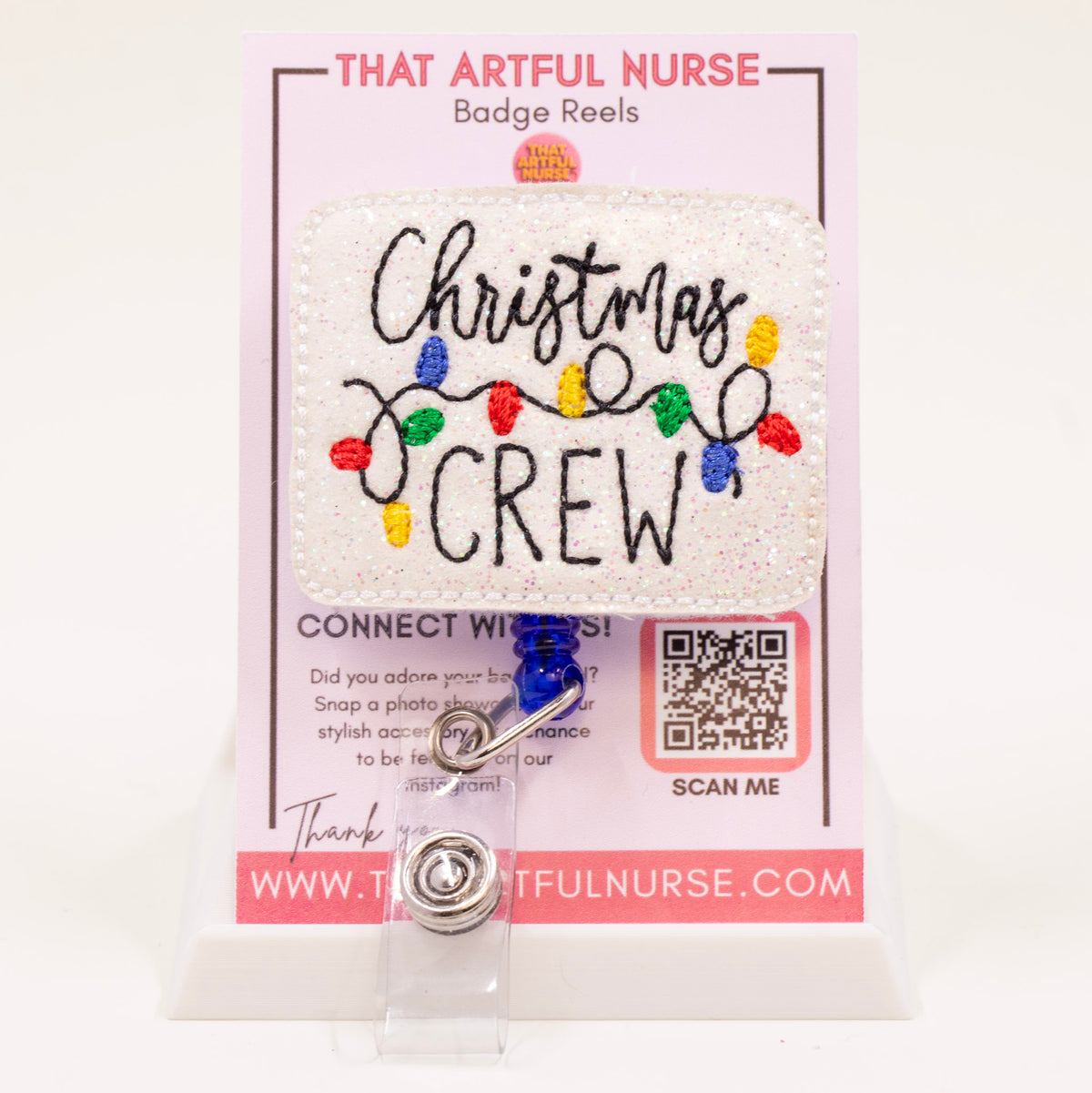 Christmas Crew badge reel with glittery white background and colorful holiday lights embroidery, displayed on That Artful Nurse packaging.