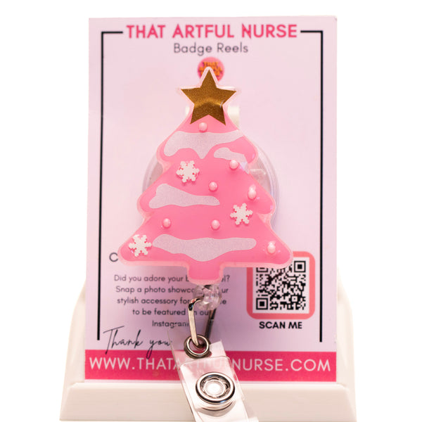 Pink Christmas tree badge reel with glitter snow accents and gold star topper, displayed on That Artful Nurse packaging.