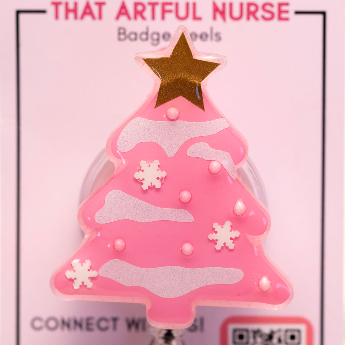 Pink Christmas tree badge reel with glitter snow accents and gold star topper, displayed on That Artful Nurse packaging.