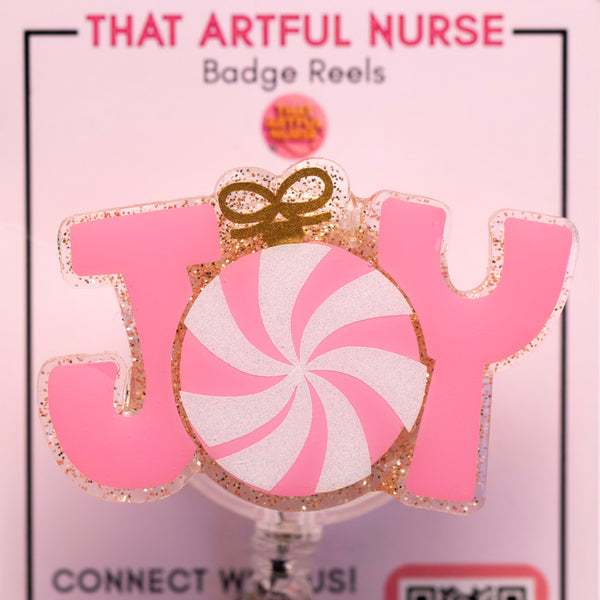 Pink JOY badge reel with glitter letters, peppermint candy center, and golden bow, displayed on That Artful Nurse packaging.