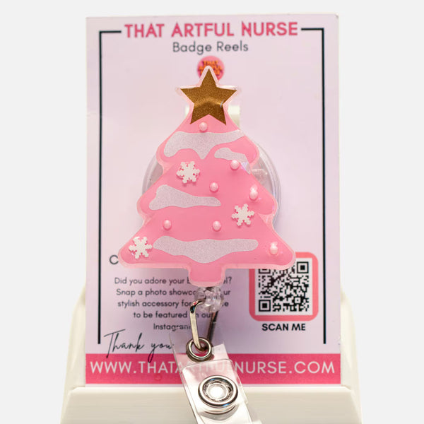 Pink Christmas tree badge reel with glitter snow accents and gold star topper, displayed on That Artful Nurse packaging.