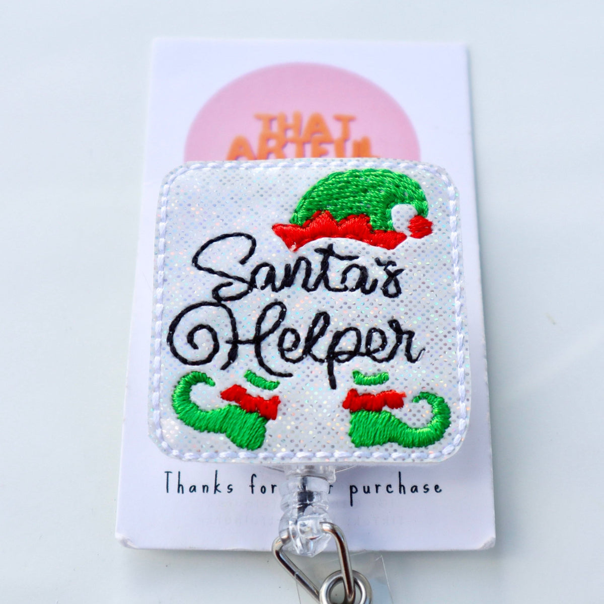 Santa’s Helper Christmas badge reel with glittery white background and embroidered elf hat and shoes.