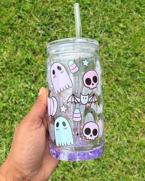Transparent tumbler featuring cute Halloween-themed designs including ghosts, skulls, bats, stars, and candy corn held by a hand against a grassy background.