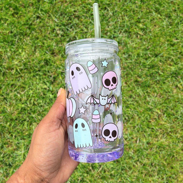 Transparent tumbler featuring cute Halloween-themed designs including ghosts, skulls, bats, stars, and candy corn held by a hand against a grassy background.
