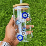 Clear glass can cup featuring a blue Turkish evil eye design held by hand against a grass background.