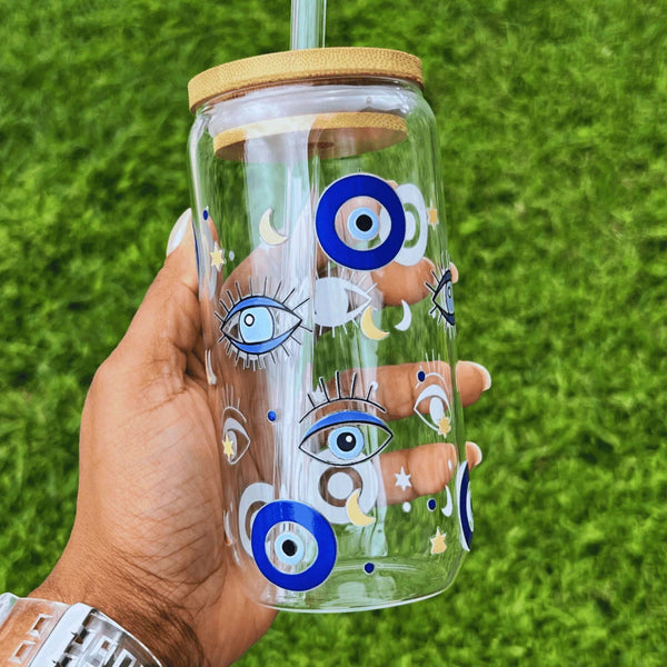 Clear glass can cup featuring a blue Turkish evil eye design held by hand against a grass background.