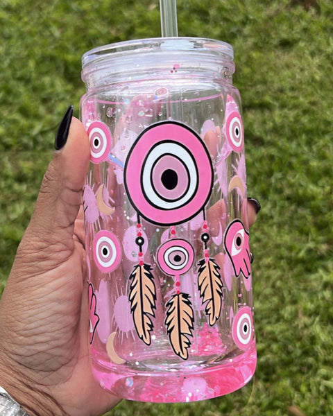 A 16oz clear snow globe tumbler with pink glitter featuring a Turkish evil eye design, held by hand on a grassy background.