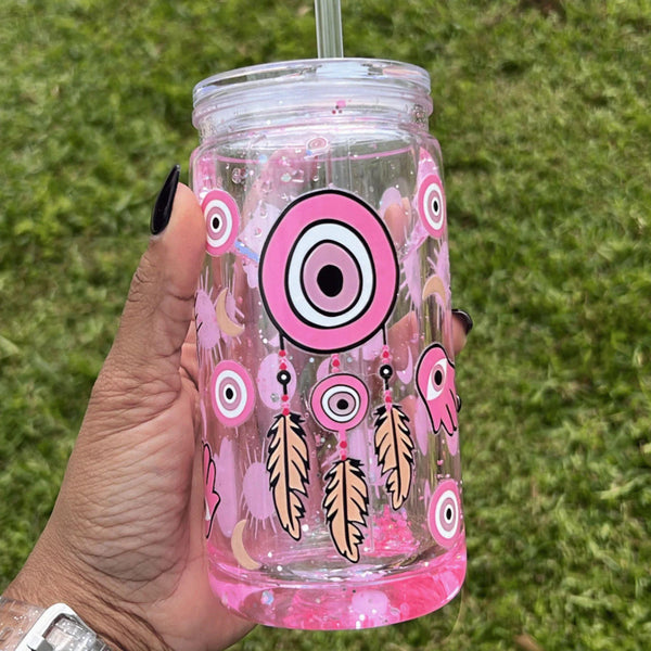 A 16oz clear snow globe tumbler with pink glitter featuring a Turkish evil eye design, held by hand on a grassy background.