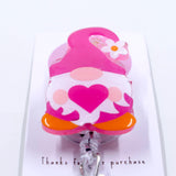 A pink gnome-shaped badge reel with a glittery heart on its outfit and a flower on its hat.