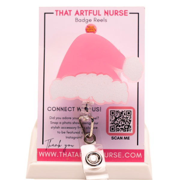 Pink Santa hat badge reel with glittery white trim on a retractable clip, displayed on That Artful Nurse packaging.