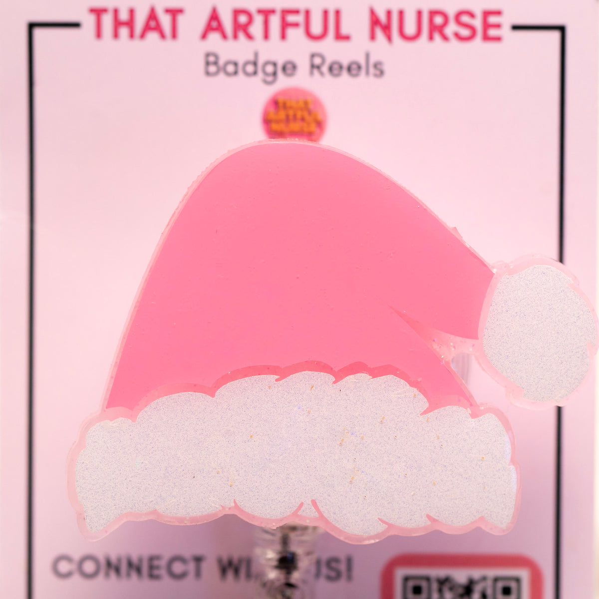 Pink Santa hat badge reel with glittery white trim on a retractable clip, displayed on That Artful Nurse packaging.