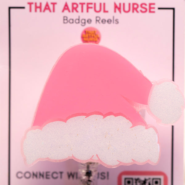 Pink Santa hat badge reel with glittery white trim on a retractable clip, displayed on That Artful Nurse packaging.